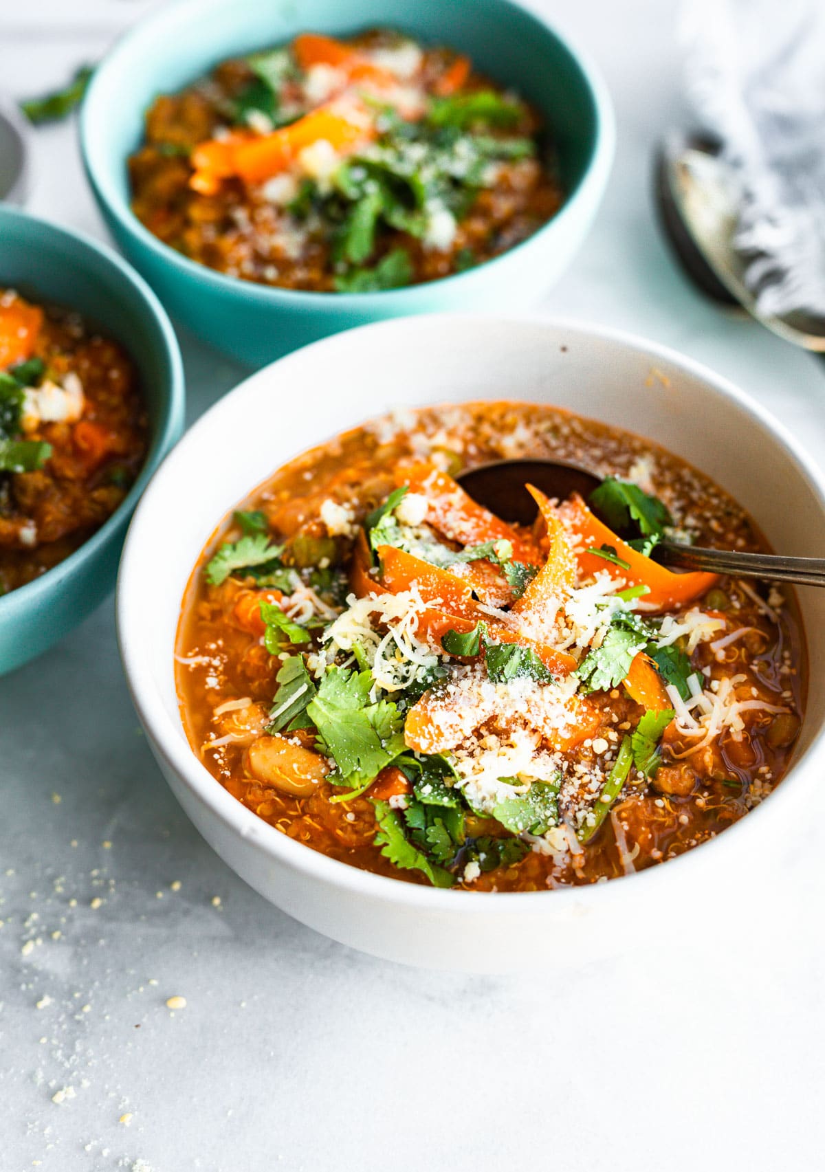 3 bowls of sausage and quinoa stew with spoon, topped with cheese, carrot shreds, and cilantro