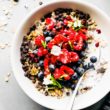 Overhead view white bowl filled with oats, quinoa, chia seeds for a breakfast bowl recipe, topped with berries and mini chocolate chips.