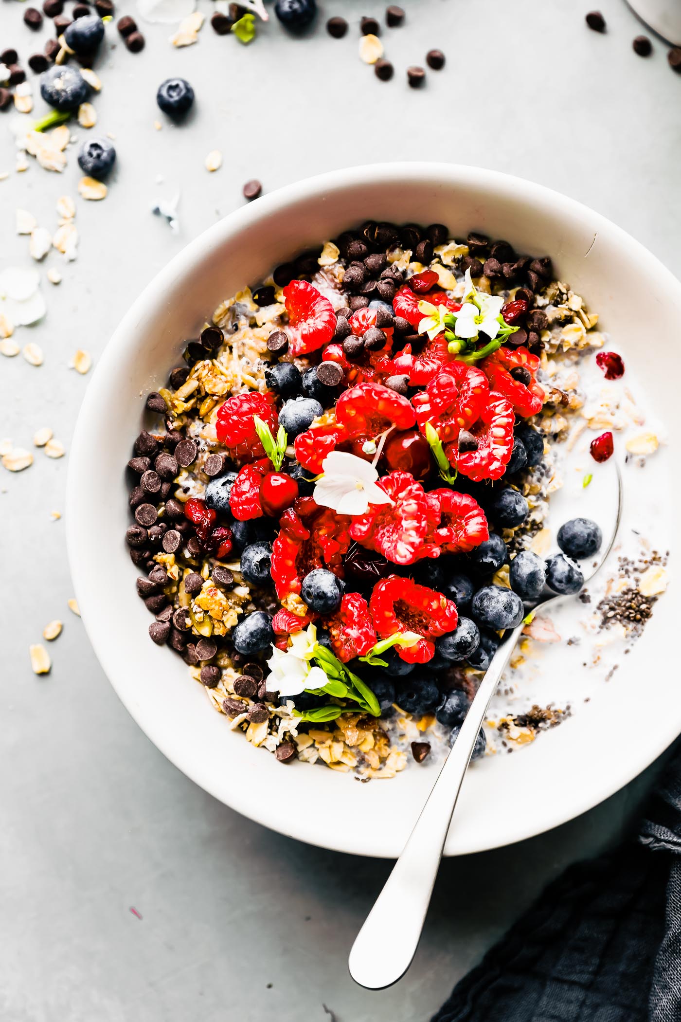 Overhead view breakfast powder bowl with grains and seeds in white bowl, topped with mini chocolate chips, fresh fruit and small flowers.