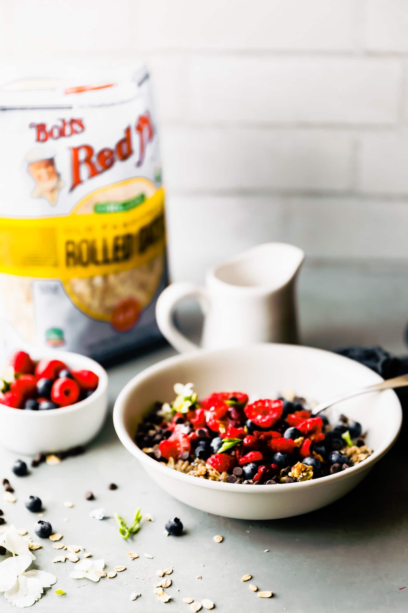 Side view bowl of breakfast power bowl filled with oats, seeds, fresh berries, and mini chocolate chips, milk pitcher and bag of oats in the background.