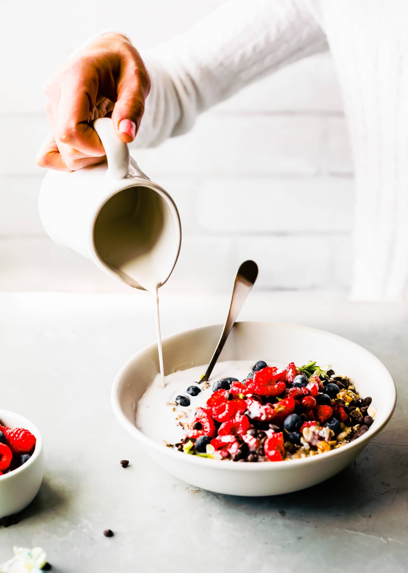 Milk from a small white pitcher being poured into a white bowl filled with oats, quinoa, chia seeds for a breakfast bowl, topped with berries and chocolate chips.
