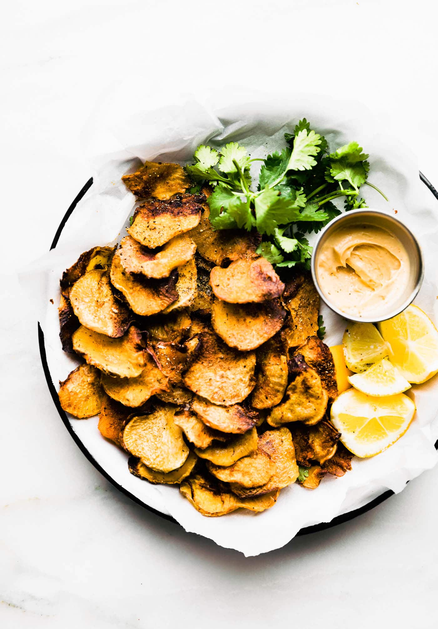 Overhead view bbq baked rutabaga chips on parchment lined round tray with dipping sauce on side.