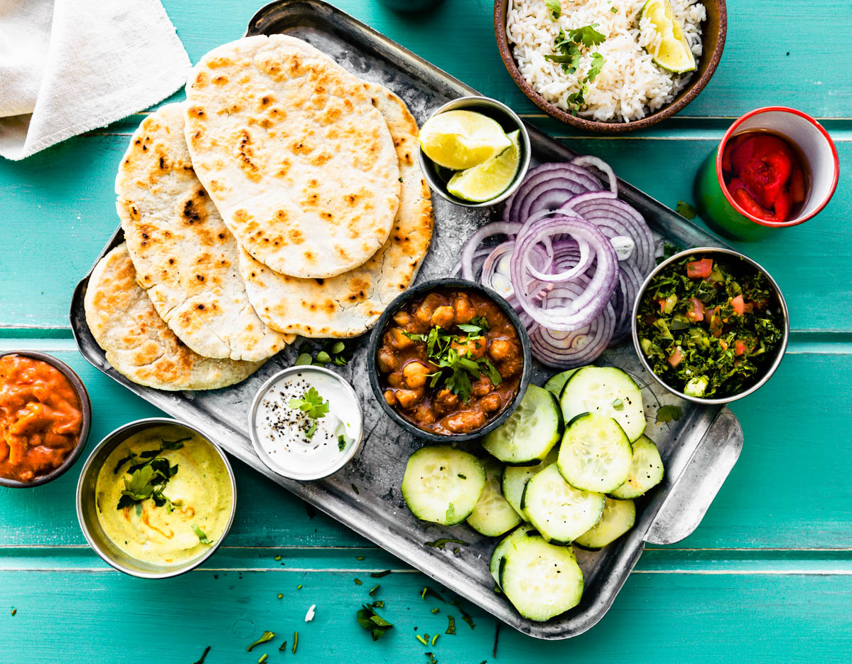 On a turquoise background, a silver baking sheet filled with several grain free naan bread and topping fixings in small bowls.