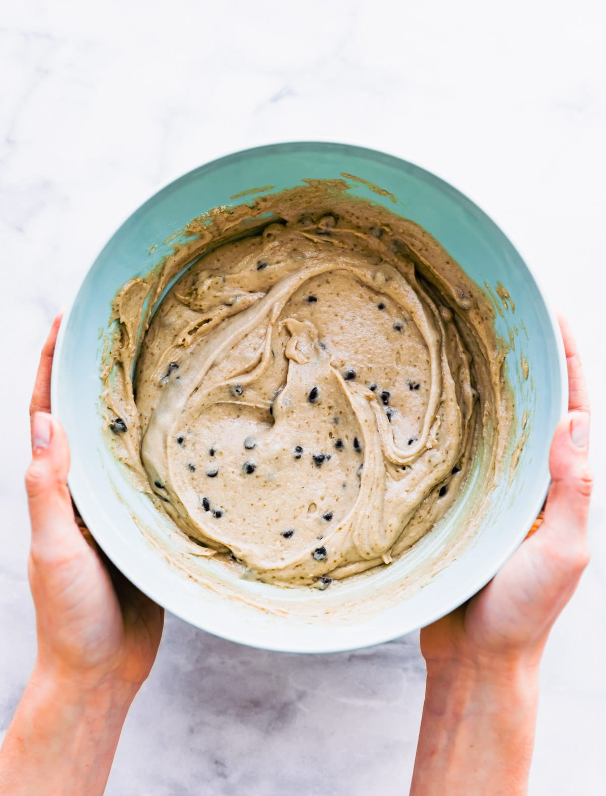 Two hands holding blue bowl filled with banana bread batter with mini chocolate chips mixed into batter