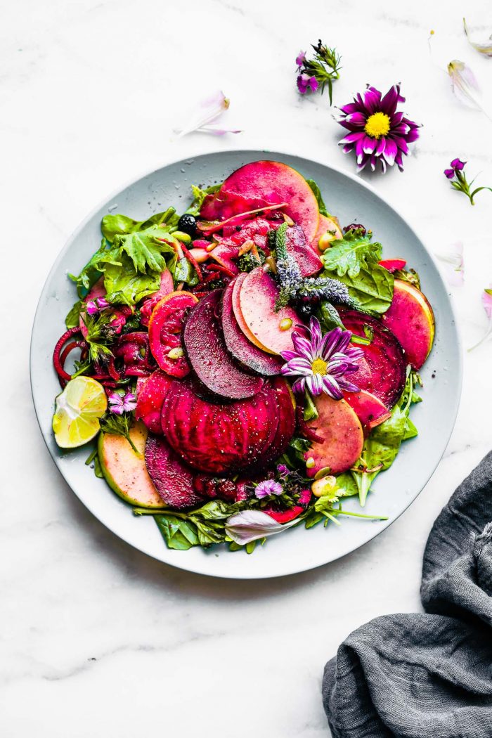 Marinated beets and apples over mixed greens on blue plate, colorful fresh cut flowers on salad.