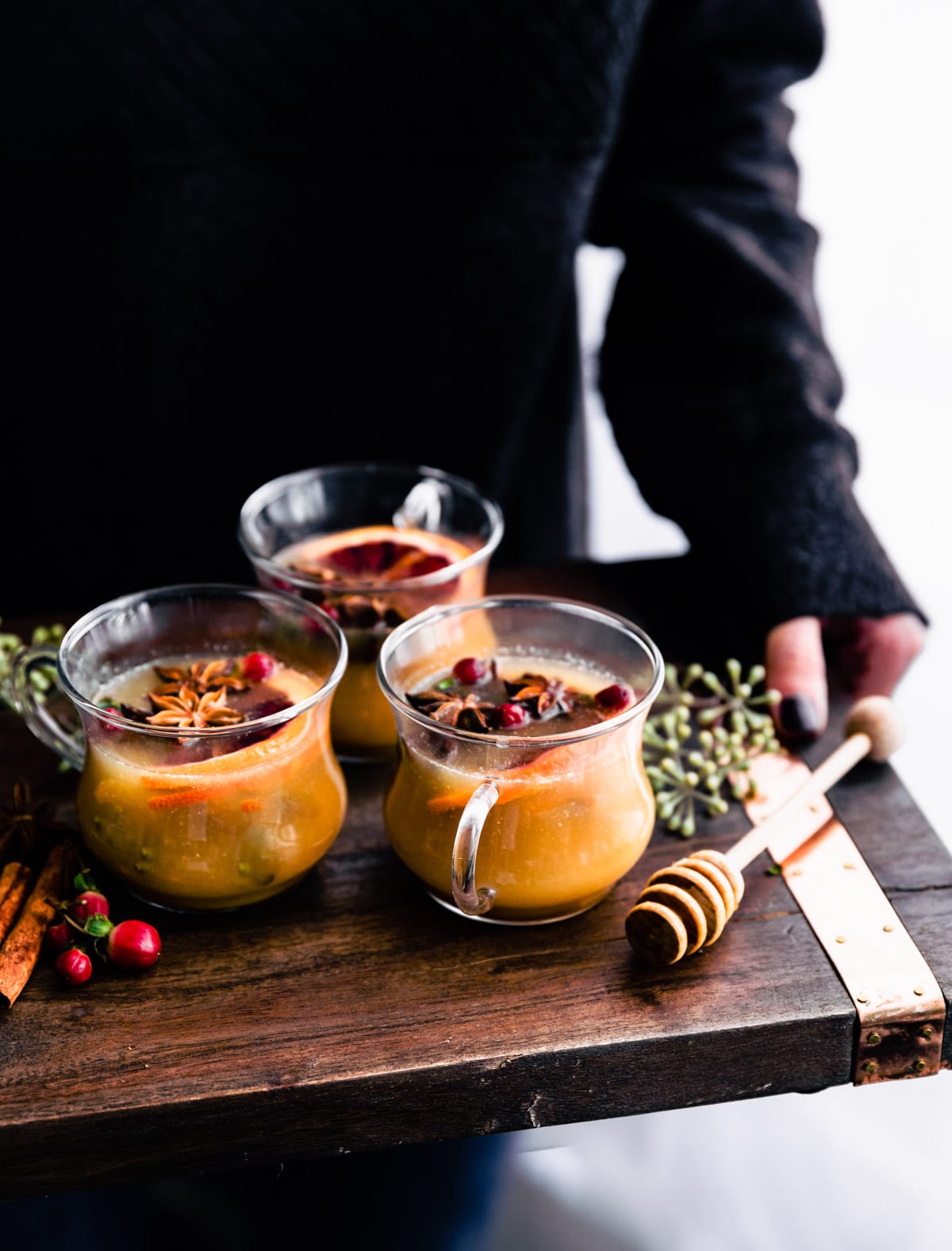 A woman holding a wooden platter holding three cups of hot toddy with spiced rum, fresh cranberries, orange slices and star anise floating in drinks