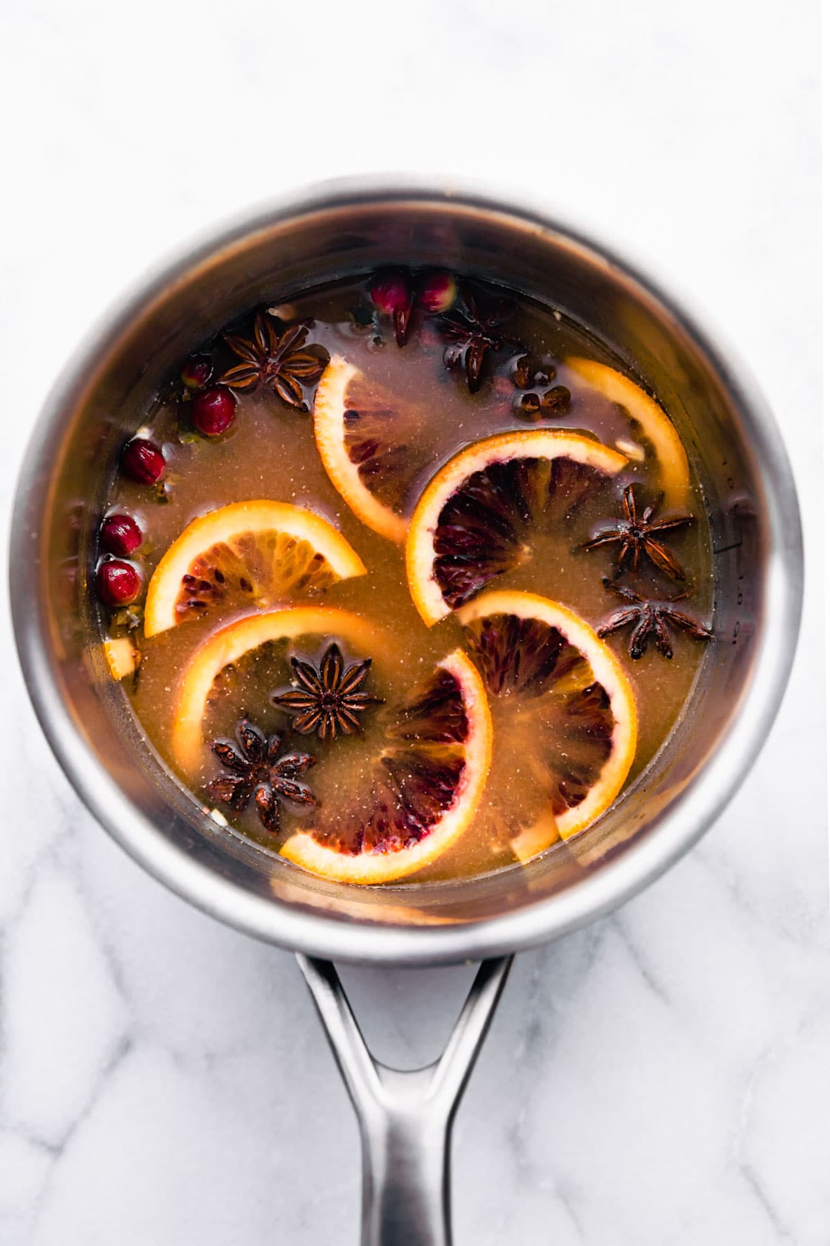 Overhead view hot toddy with spiced rum in silver saucepan with fresh cranberries, orange slices, and star anise floating in drink.