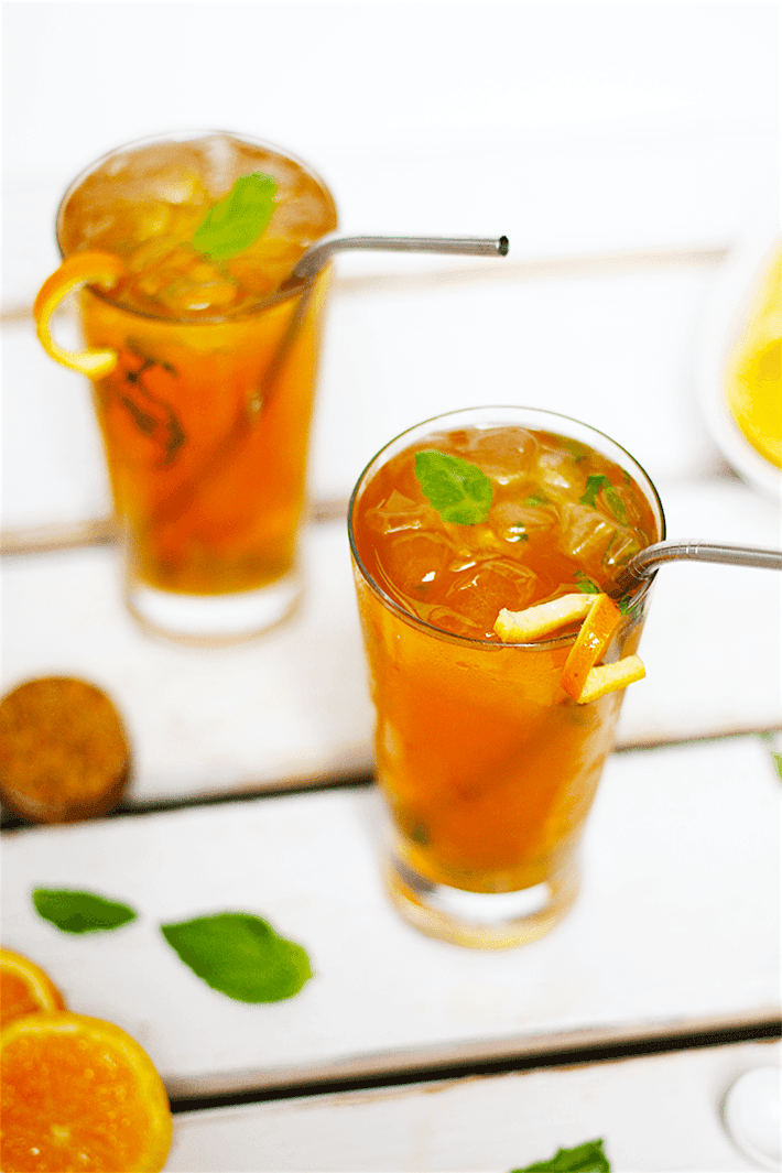 Close up view of two tall glasses of orange and basil adrenal cocktail; metal straw in glasses and garnished with orange rind twist and fresh basil.