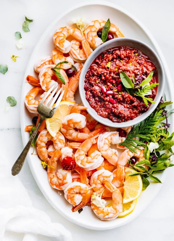 Overhead view of shrimp served on a white platter with cranberry basil cocktail dipping sauce in a white bowl.