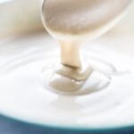 Smooth white cashew cream sauce pouring off a spoon into a blue bowl