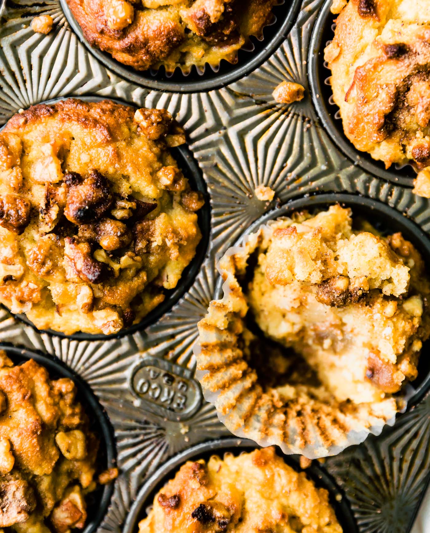Overhead view muffin tin with spiced pear muffins with nut toppings. One muffin with bite taken out placed sideways in tin to show inside texture.