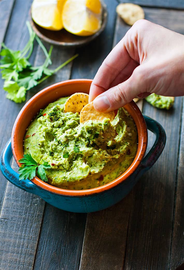 Easy Chimichurri White Bean Hummus in an orange and blue bowl with two round crackers being dipped into the creamy hummus.