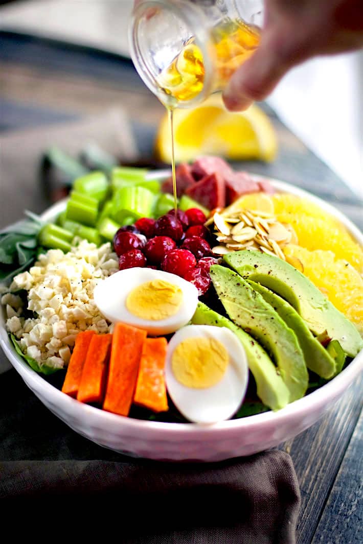 Homemade vinaigrette being drizzled on top of a winter seasonal Cobb Salad topped with fresh cranberries, almonds, avocado, hard boiled egg, and carrots; served in a white bowl