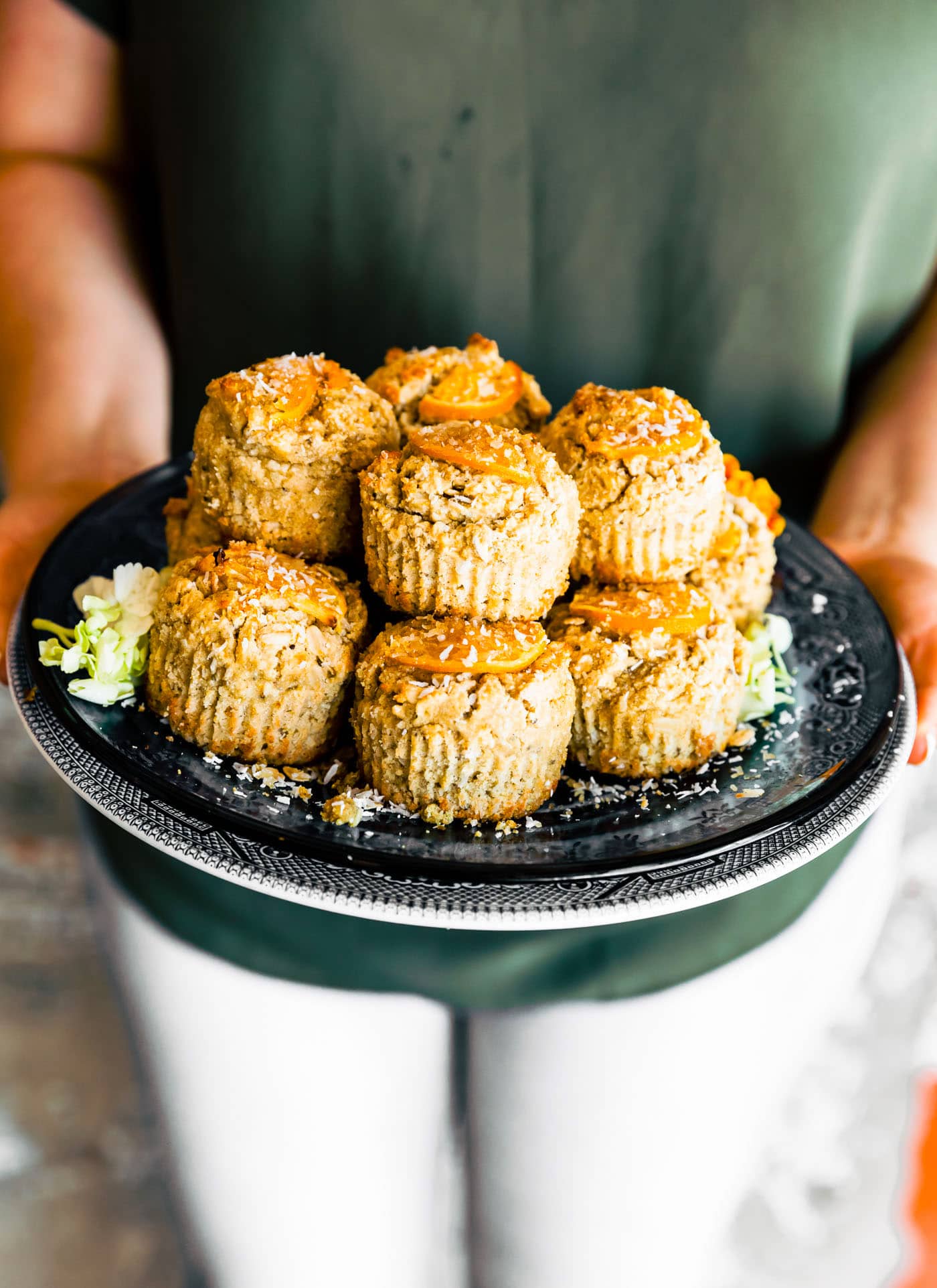 Orange oat muffins stacked up on a black tray being held by a woman wearing green.