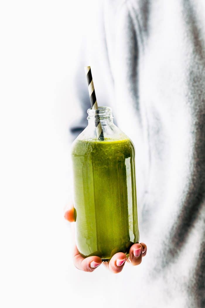 A hand holding a glass bottle of green smoothie with black and white straw in smoothie.
