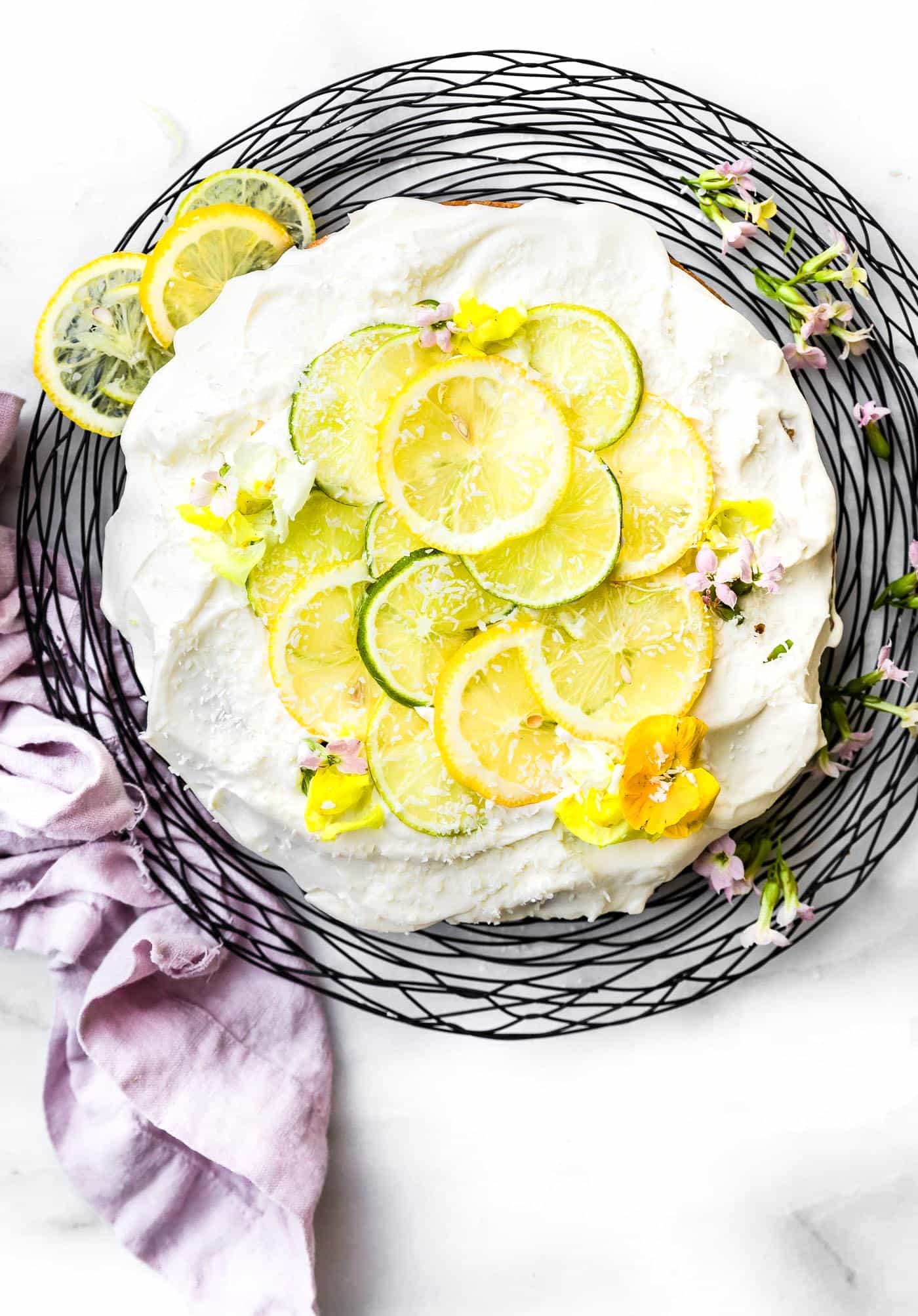 Overhead view coconut vegan cake topped with white frosting and thin slices lemon and lime, on round black wire cake stand.