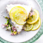Overhead view citrus coconut vegan cake with white frosting, lemon slices, and coconut shreds on white plate.