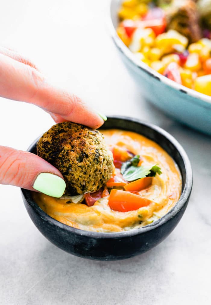 A hand holding a Mexican vegan falafel ball dipping it into an orange sauce in a black bowl.