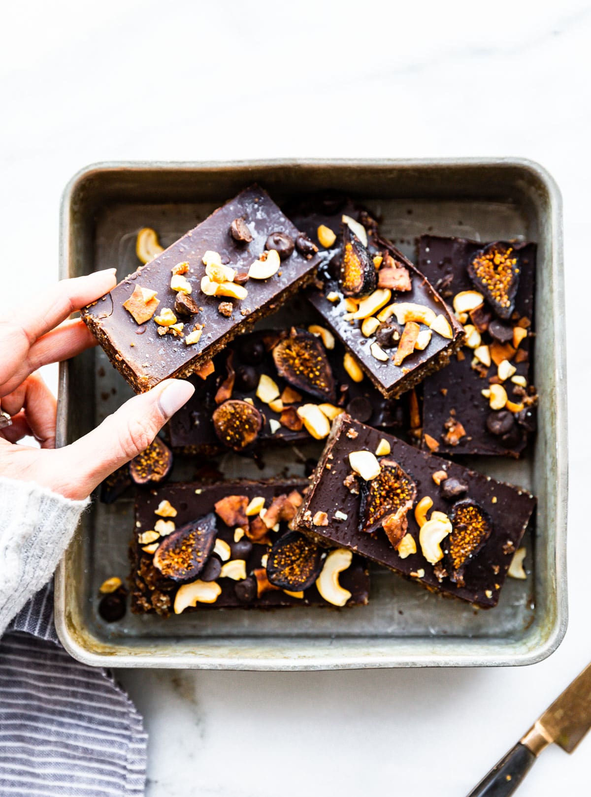 No Bake Chocolate Coconut Cashew Fig Bars cut into long bars in square baking sheet, a hand holding up one bar