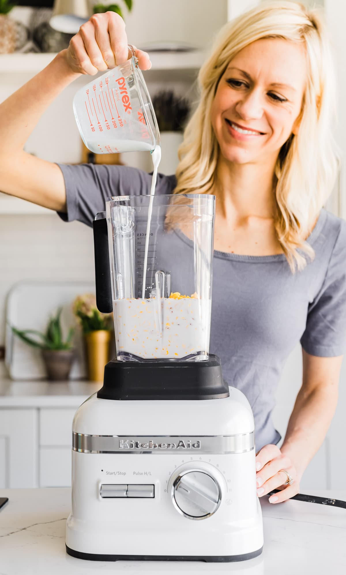 A woman standing behind kitchenaid blender pouring milk into blender cup.