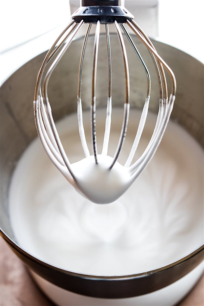 Whipped aquafaba on a stand mixer whisk hovering above a silver mixing bowl filled with easy vegan aquafaba.