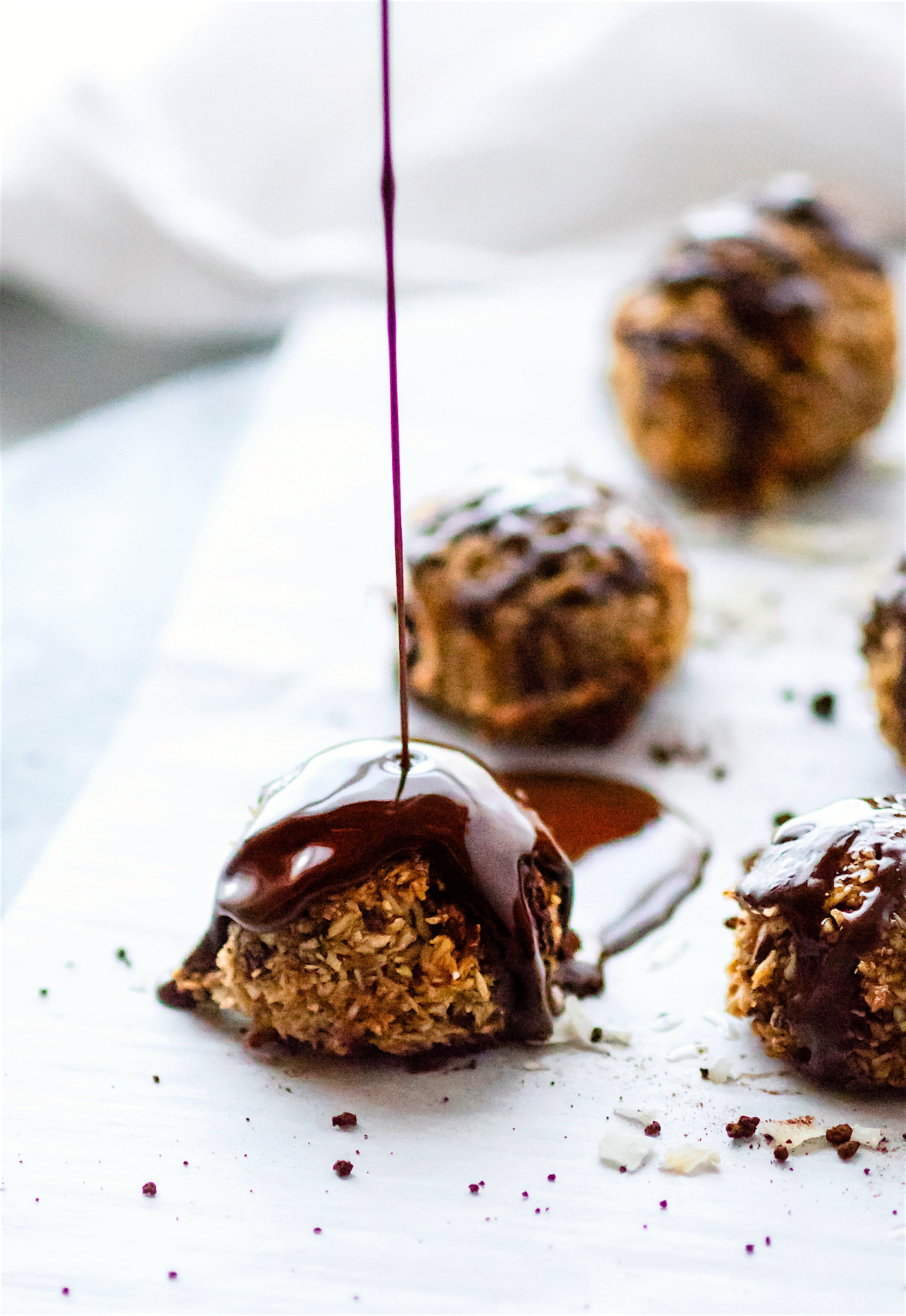 A thick drizzle of melted dark chocolate being poured over a coconut macaroon on a sheet of parchment paper