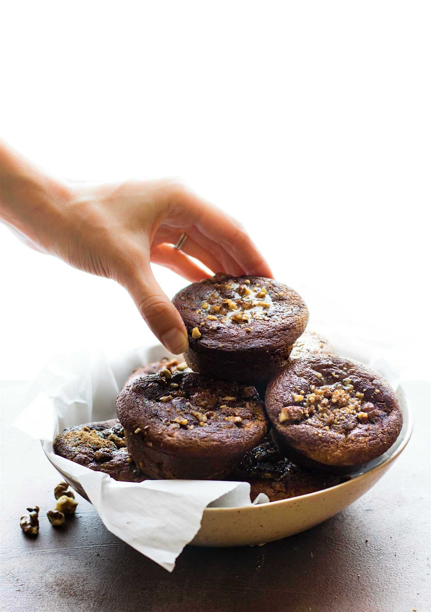 A coffee cake muffin with sticky nut topping being picked up from a serving bowl filled with gluten-free muffins.