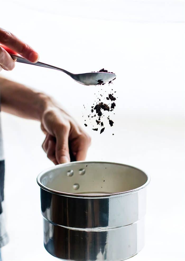 Coffee grounds being sprinkled into a saucepan being held by a hand.