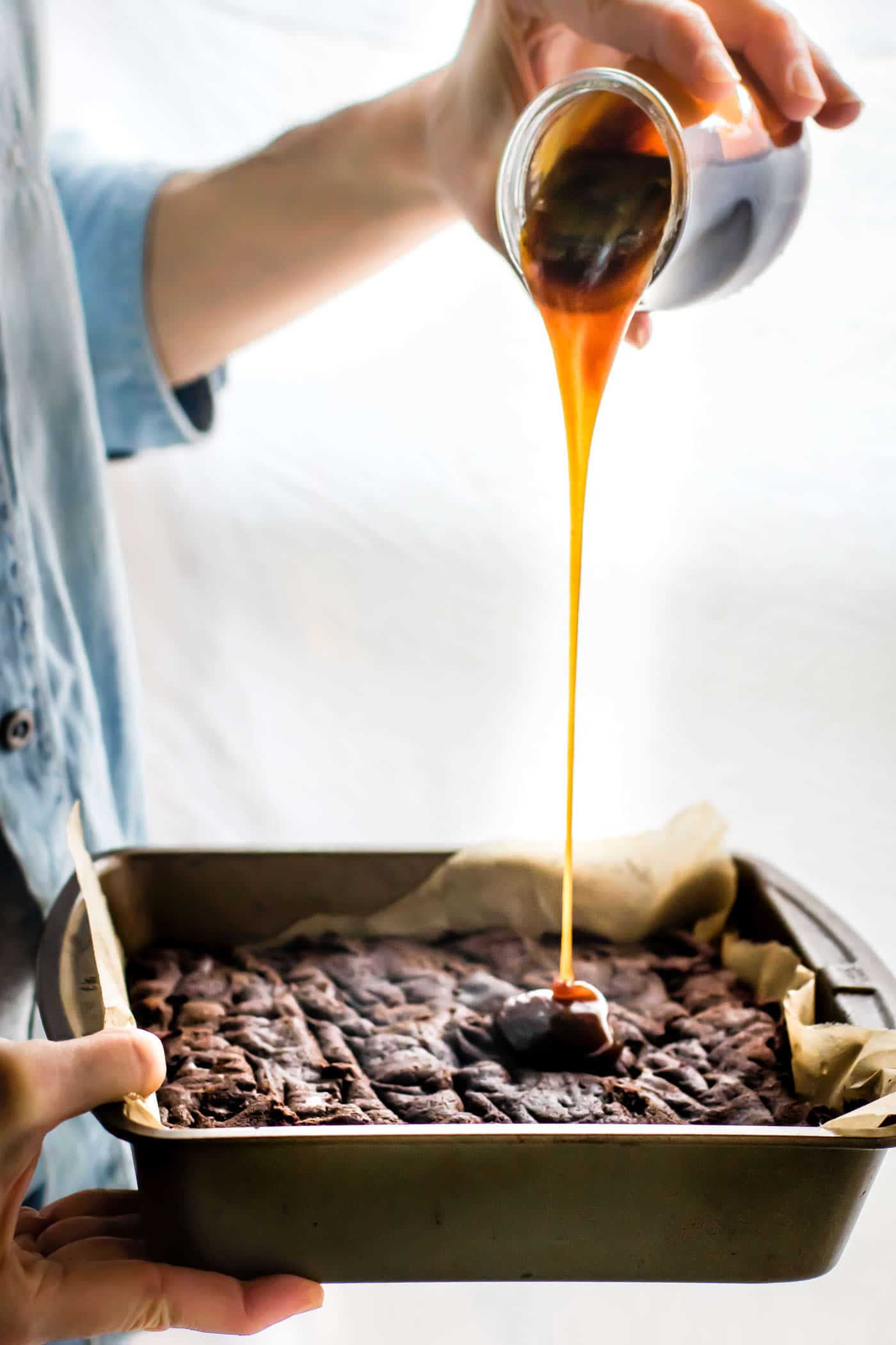 Caramel sauce being drizzled from a glass jar onto a pan of dark chocolate gluten-free brownies.