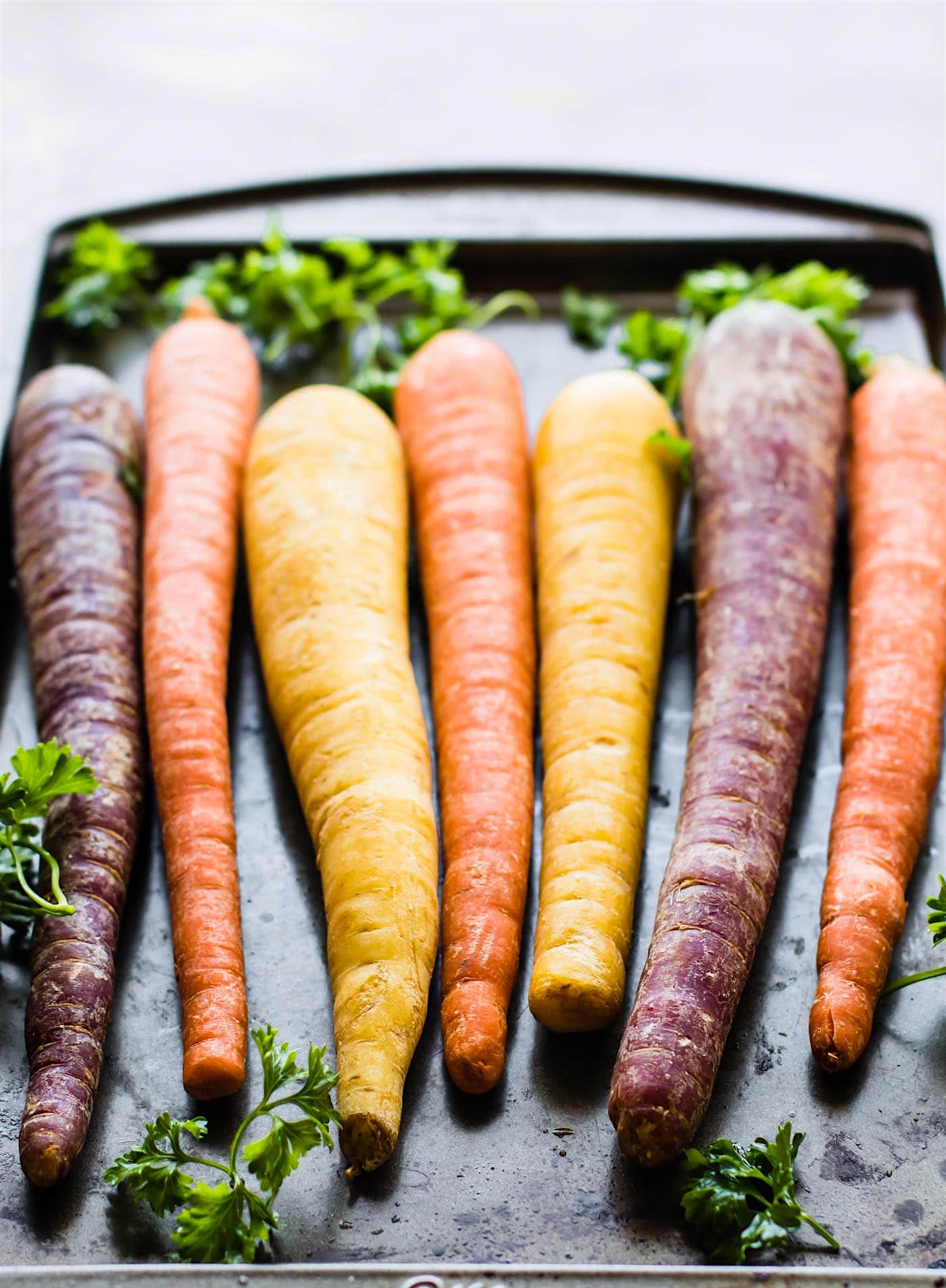 Several rainbow colored carrots with green leaved tops laying on a baking sheet.