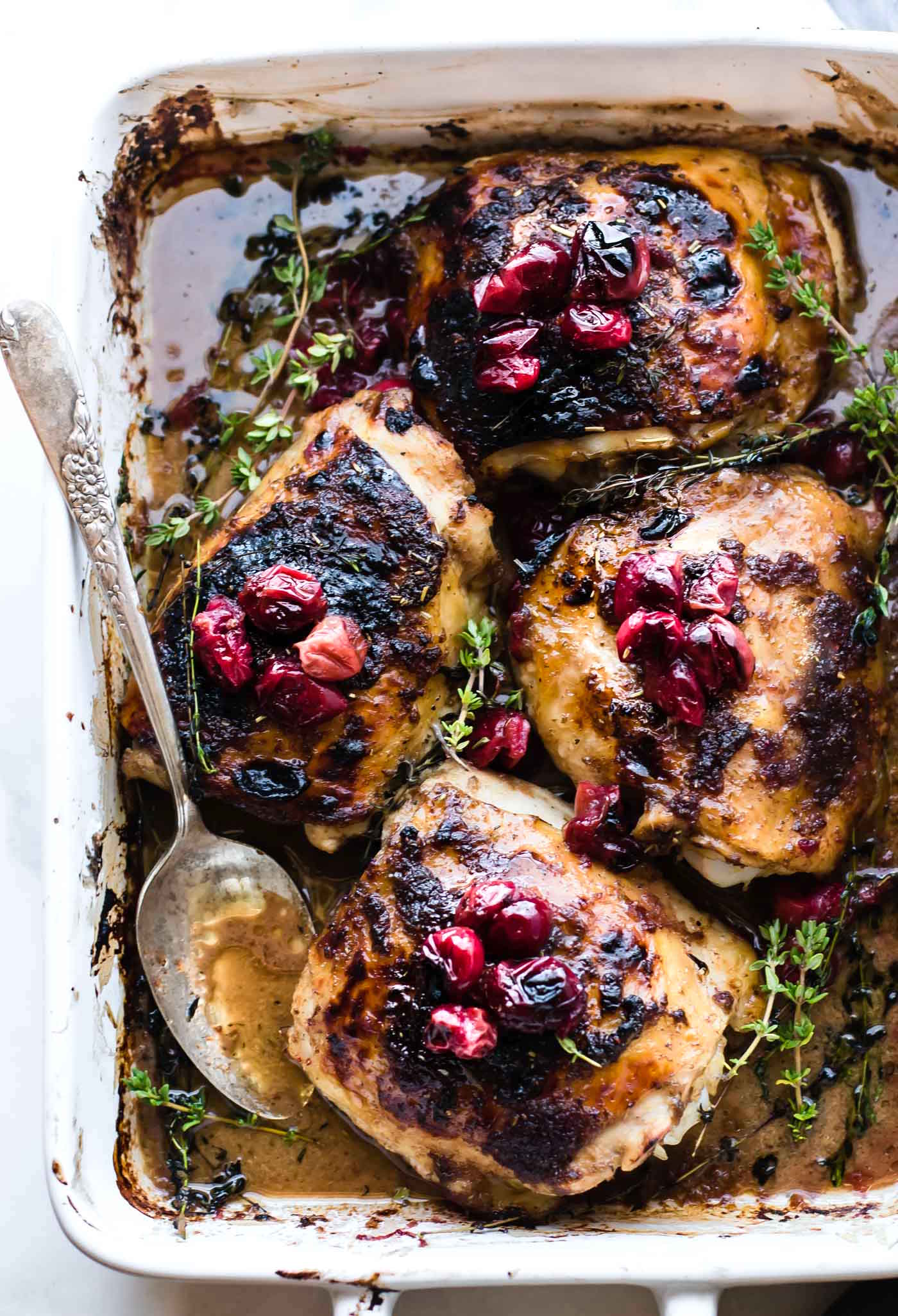 overhead photo: slow roasted cranberry balsamic chicken in a white baking pan with serving spoon