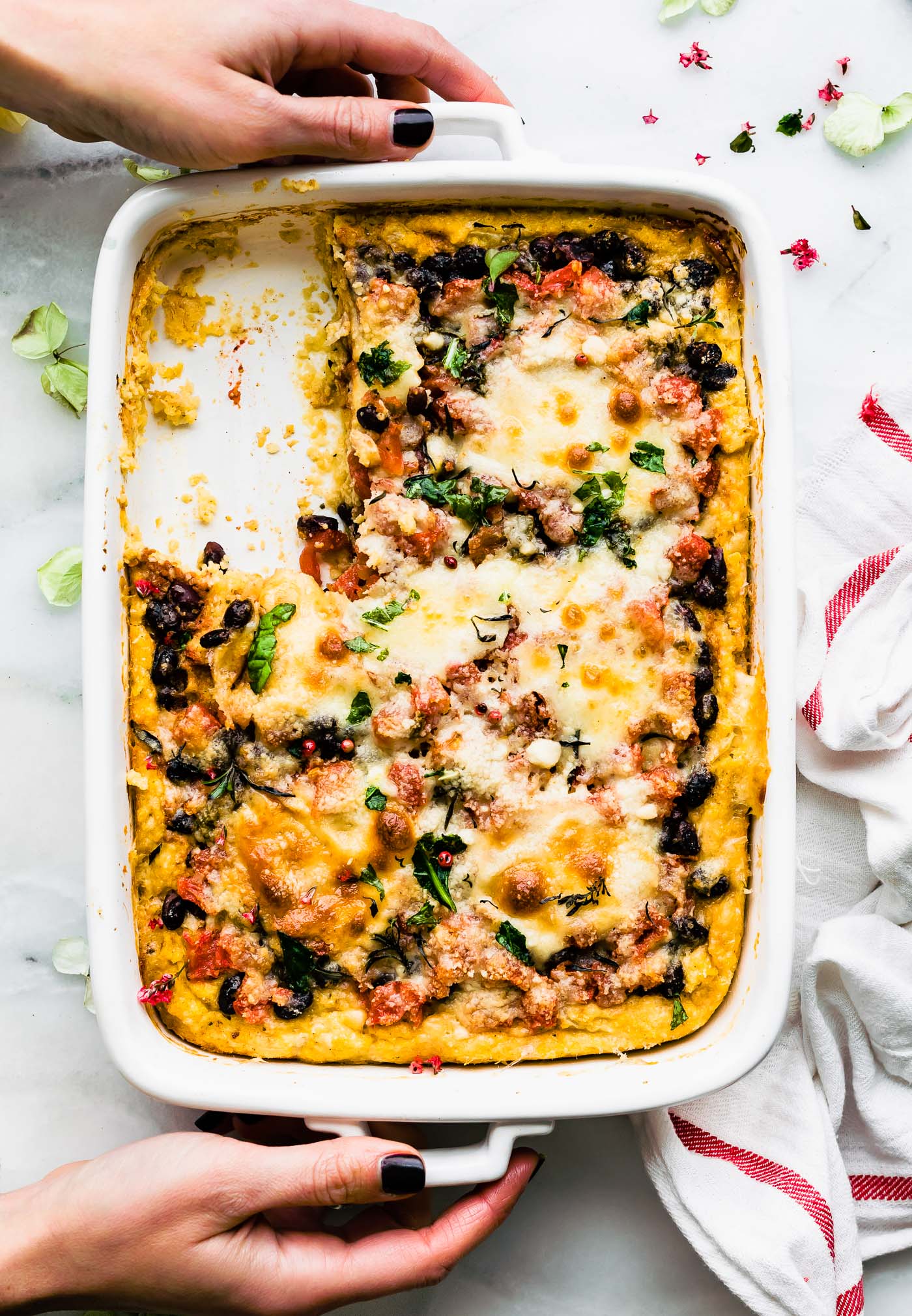 two hands holding a casserole dish of black bean & polenta casserole with a serving mising