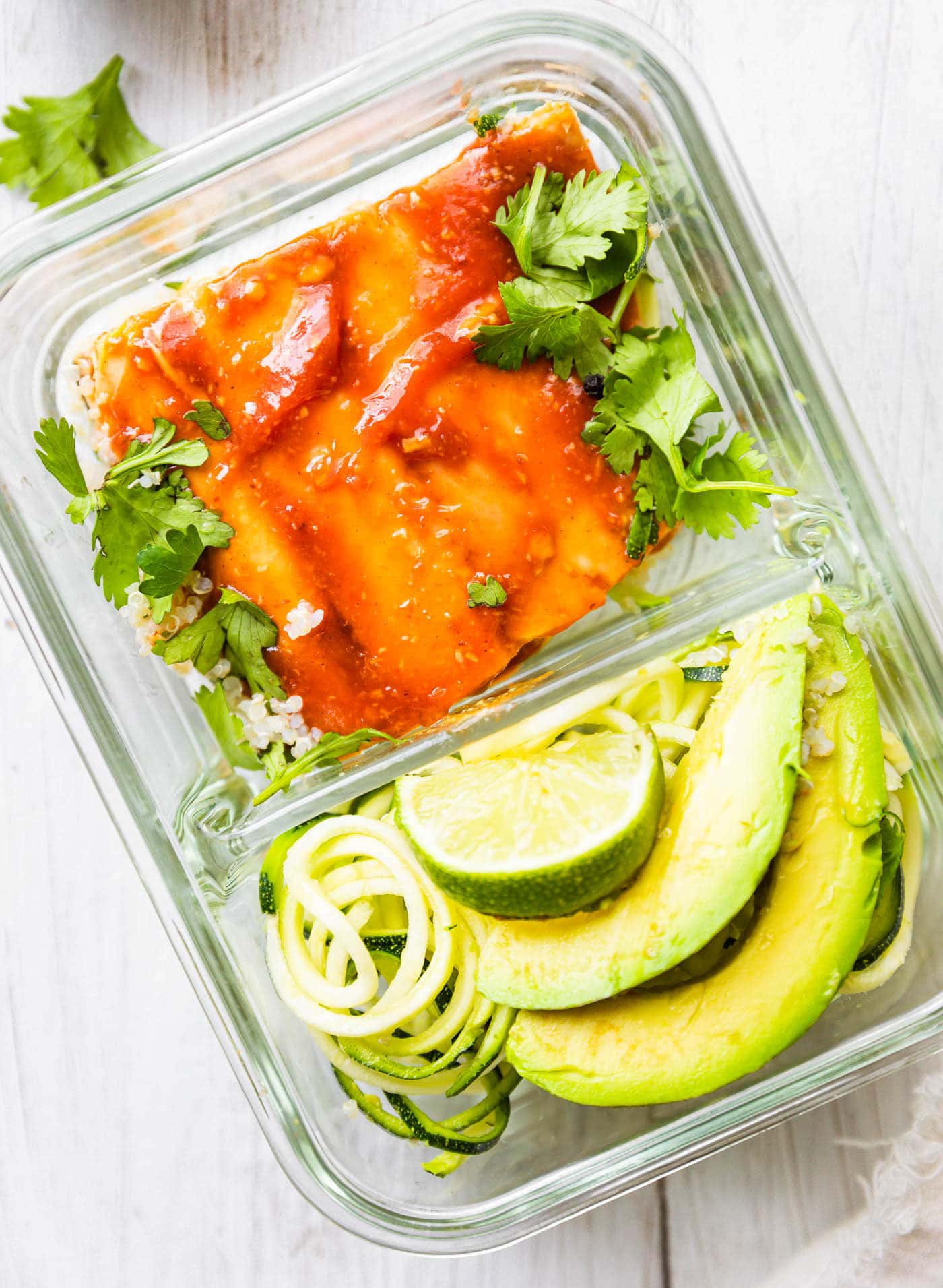 Glass divided meal prep container with bbq salmon fillet in one side and zucchini spirals and avocado slices in other side.