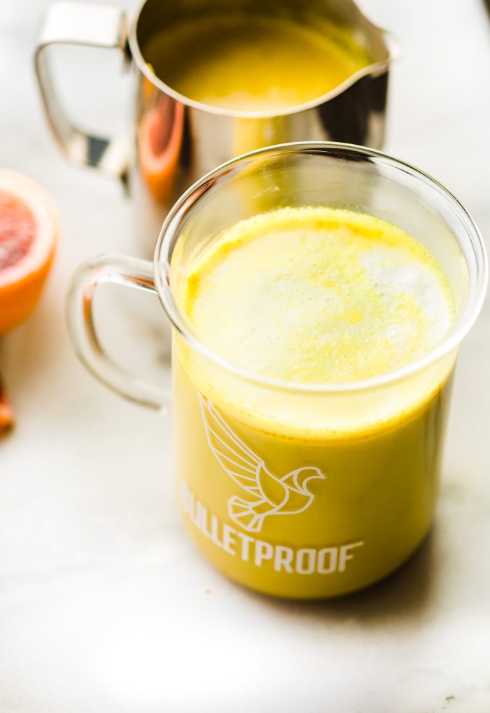 Orange turmeric latte in clear glass mug and in background, metal frothing pitcher filled with latte