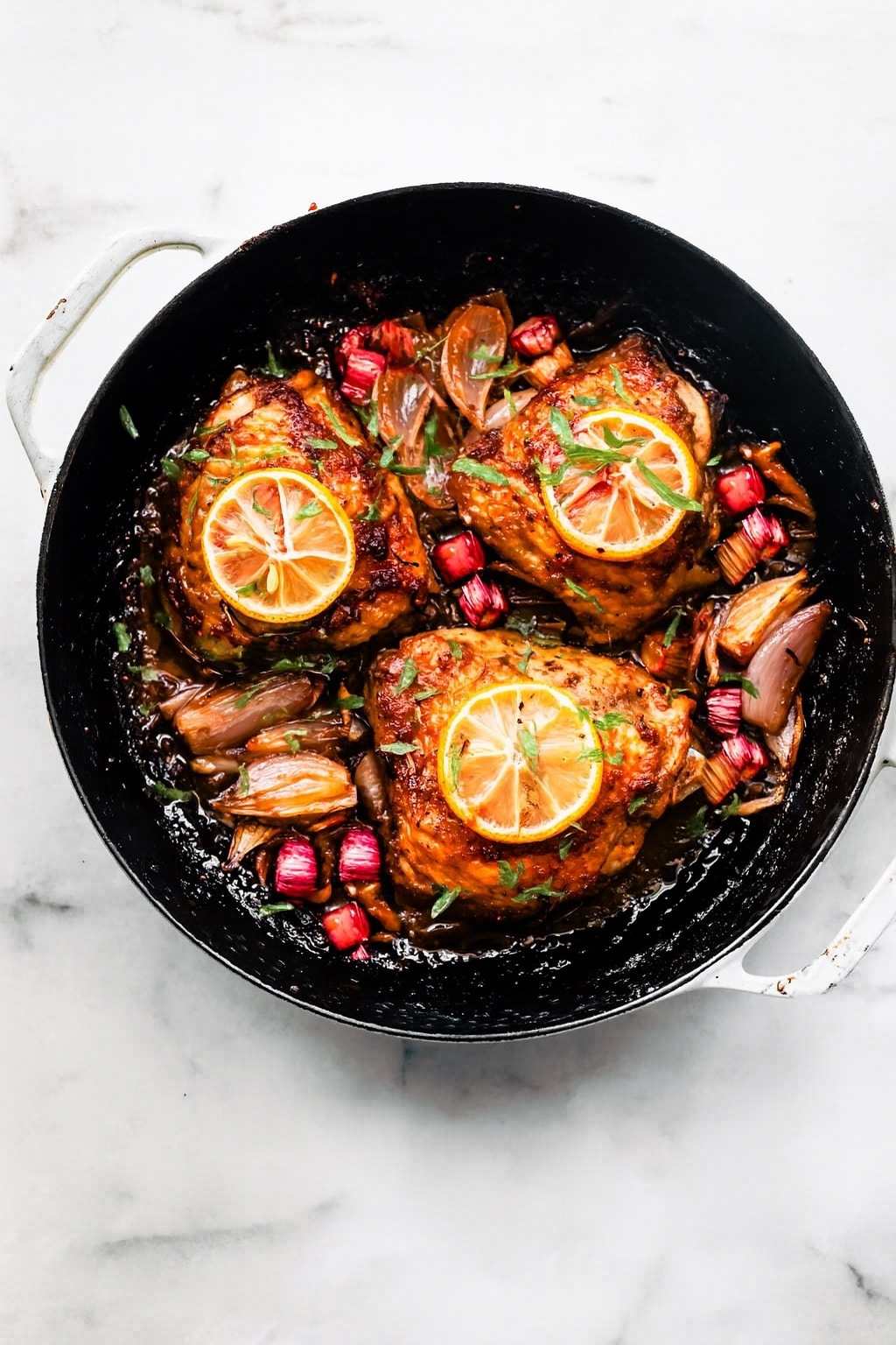 Roasted chicken thighs in a pan with caramelized shallots, chunks of rhubarb, and lemon slices on top.