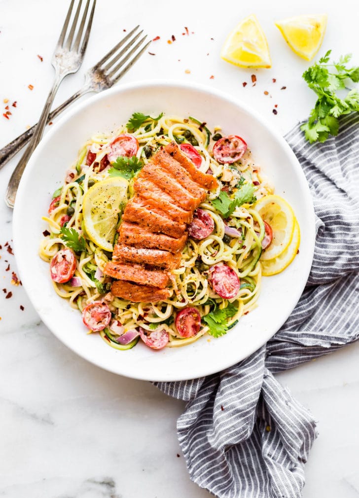 Smoked Salmon on a bed of zucchini noodles with sauce and cherry tomatoes, plated on a white plate with two forks next to plate.
