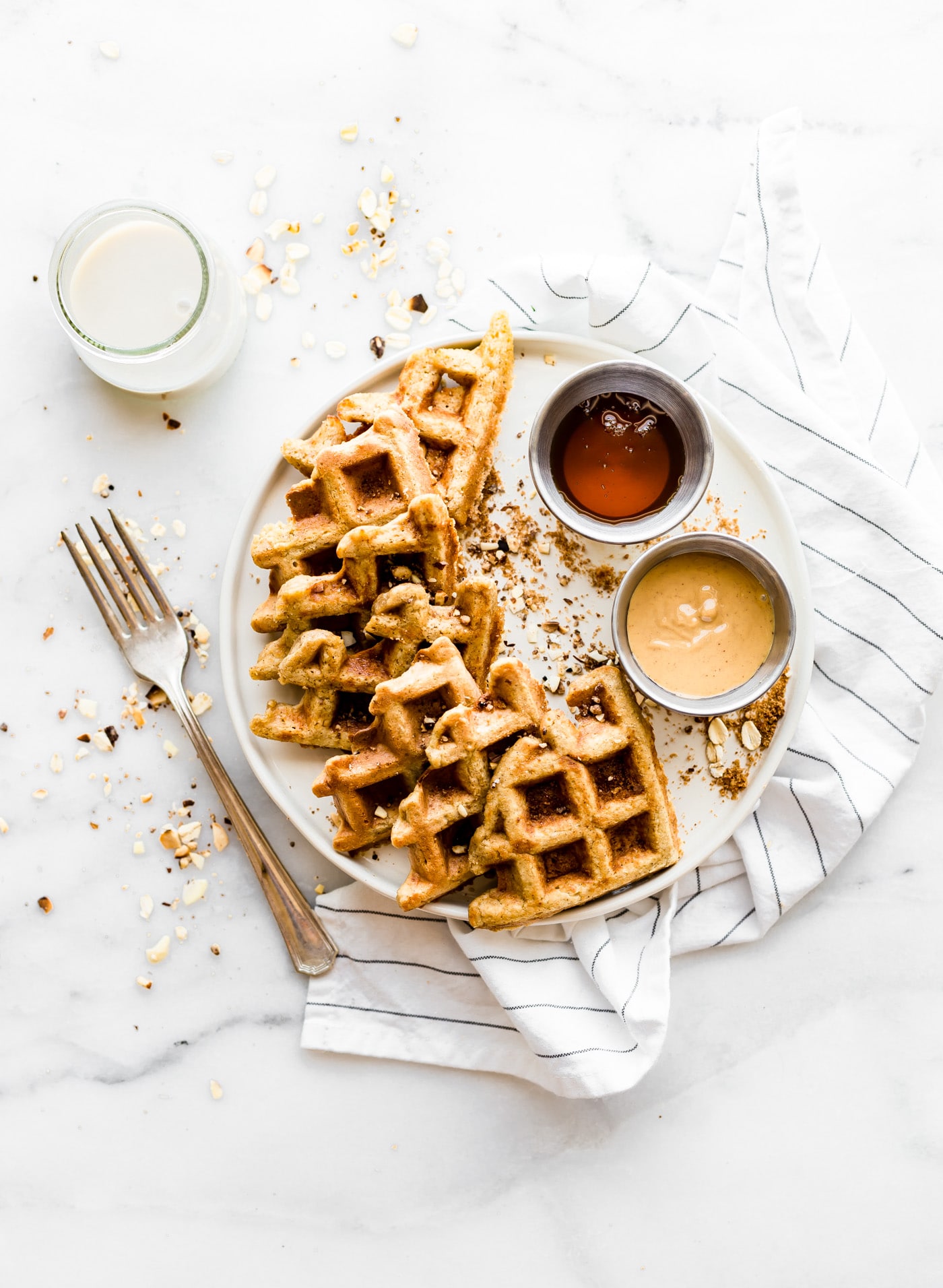 Several flourless peanut butter waffles on white plate with peanut butter on side and maple syrup on side.