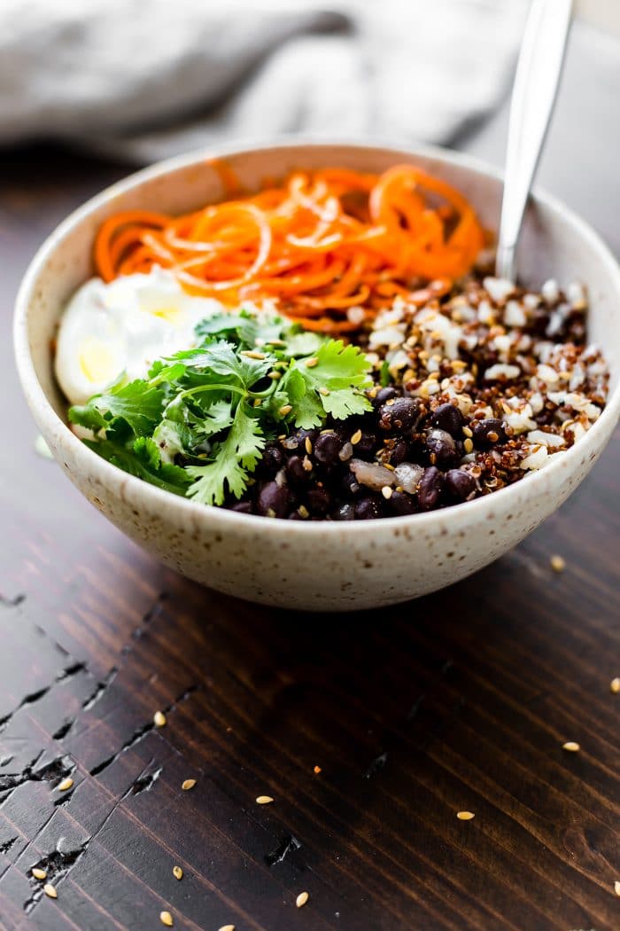 A cream colored bowl filled with grains, black beans, fresh cilantro, cream, and carrot spirals