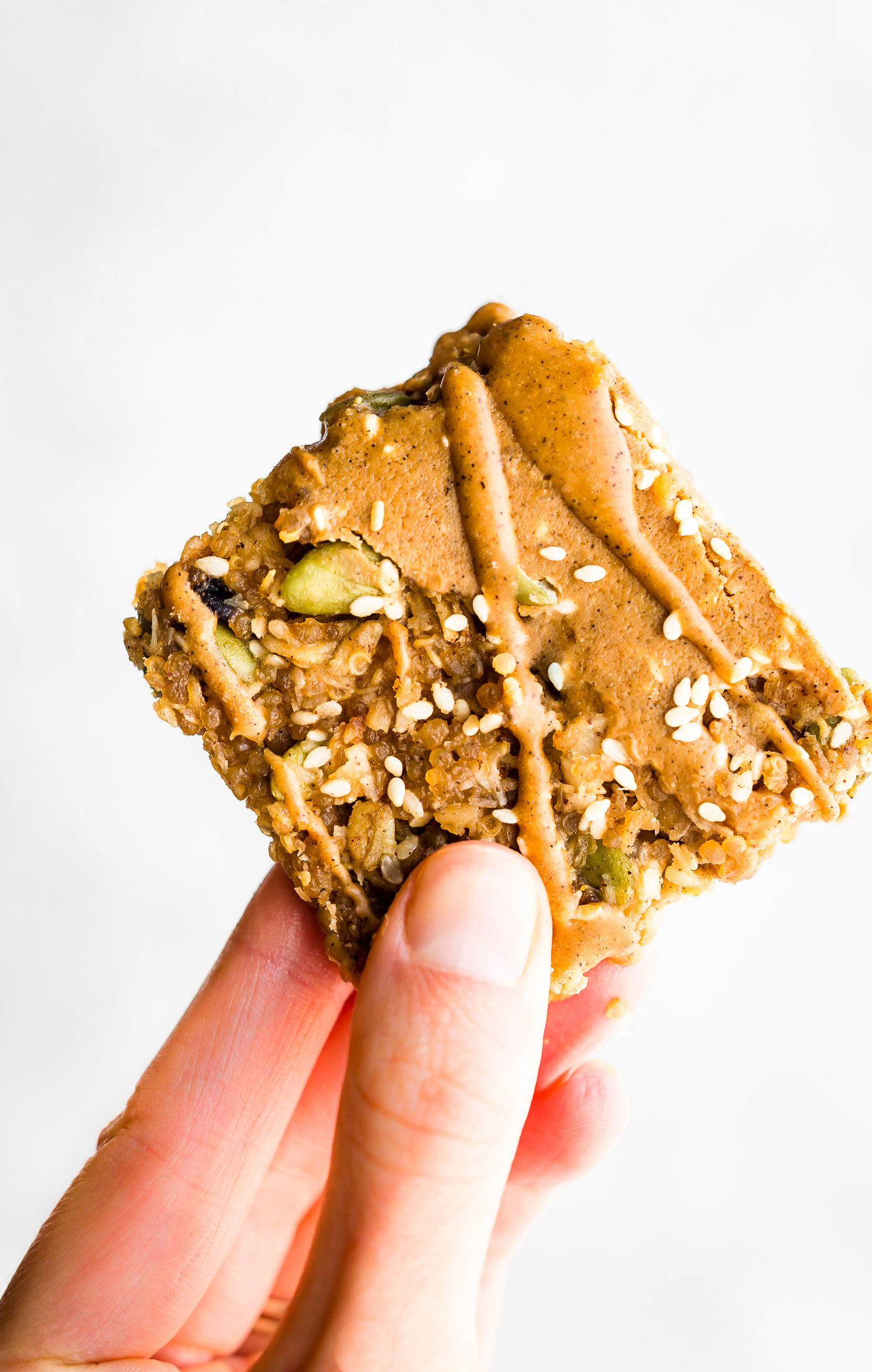 A single maple sesame quinoa bar drizzled with sunflower seed butter being held up against white background.