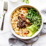 Serving of slow cooker oatmeal in stone bowl topped with crispy bacon and avocado slices.