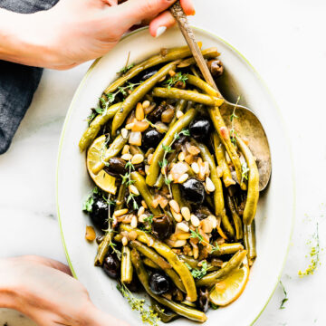 Two hands holding white oval serving try filled with balsamic olive oil braised green beans.
