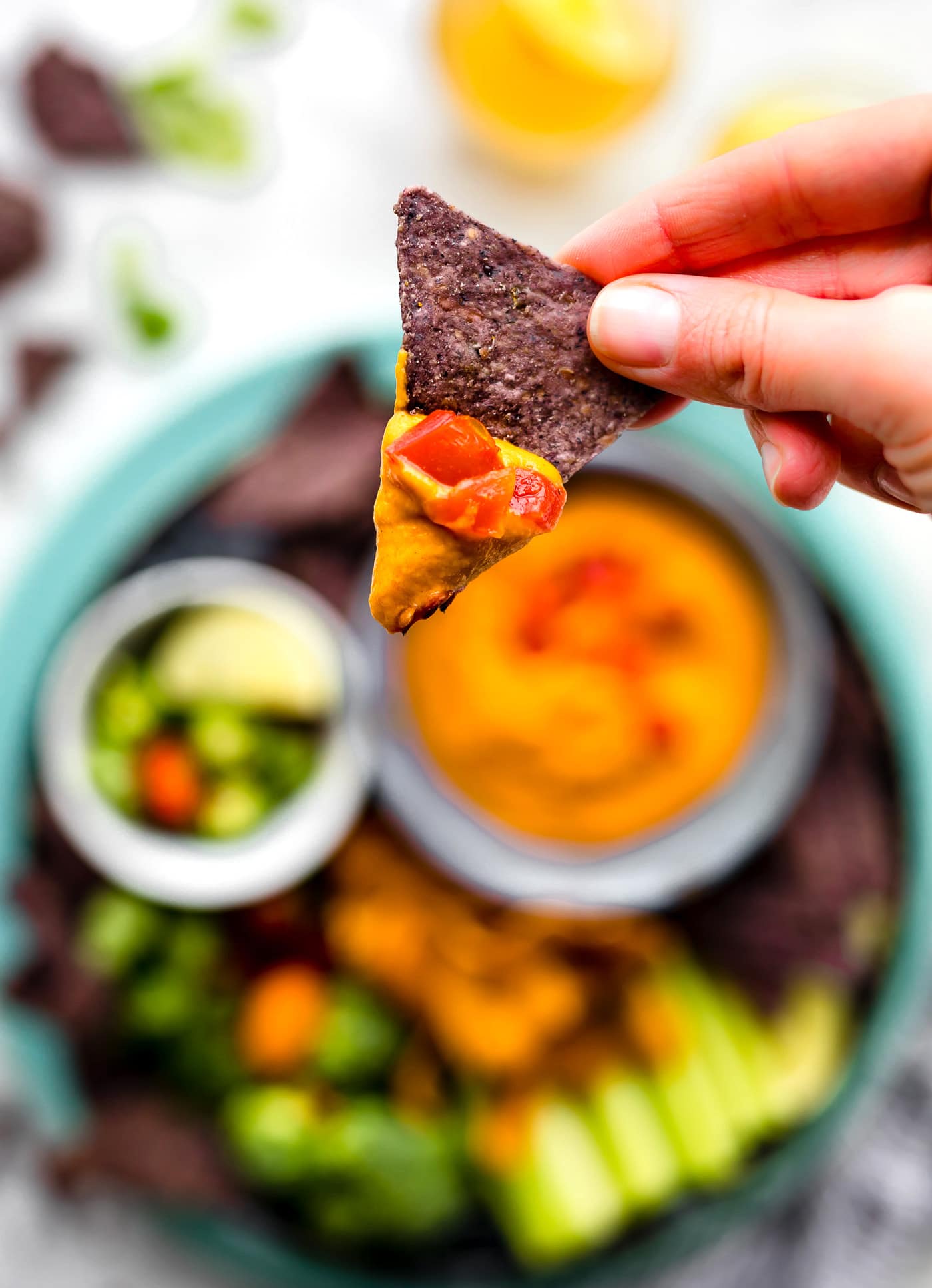 A hand holding a blue tortilla with vegan con queso on tip with tomato chunks on top of queso.