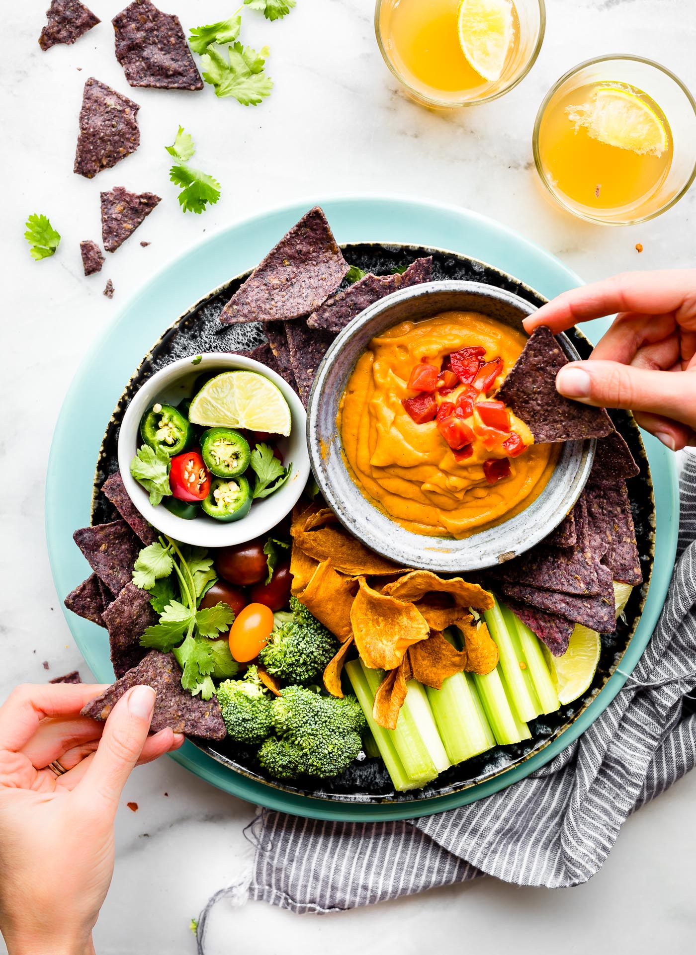 Overhead view round platter with colored tortilla chips and bowl of vegan con queso, a hand dipping a blue chip in queso.