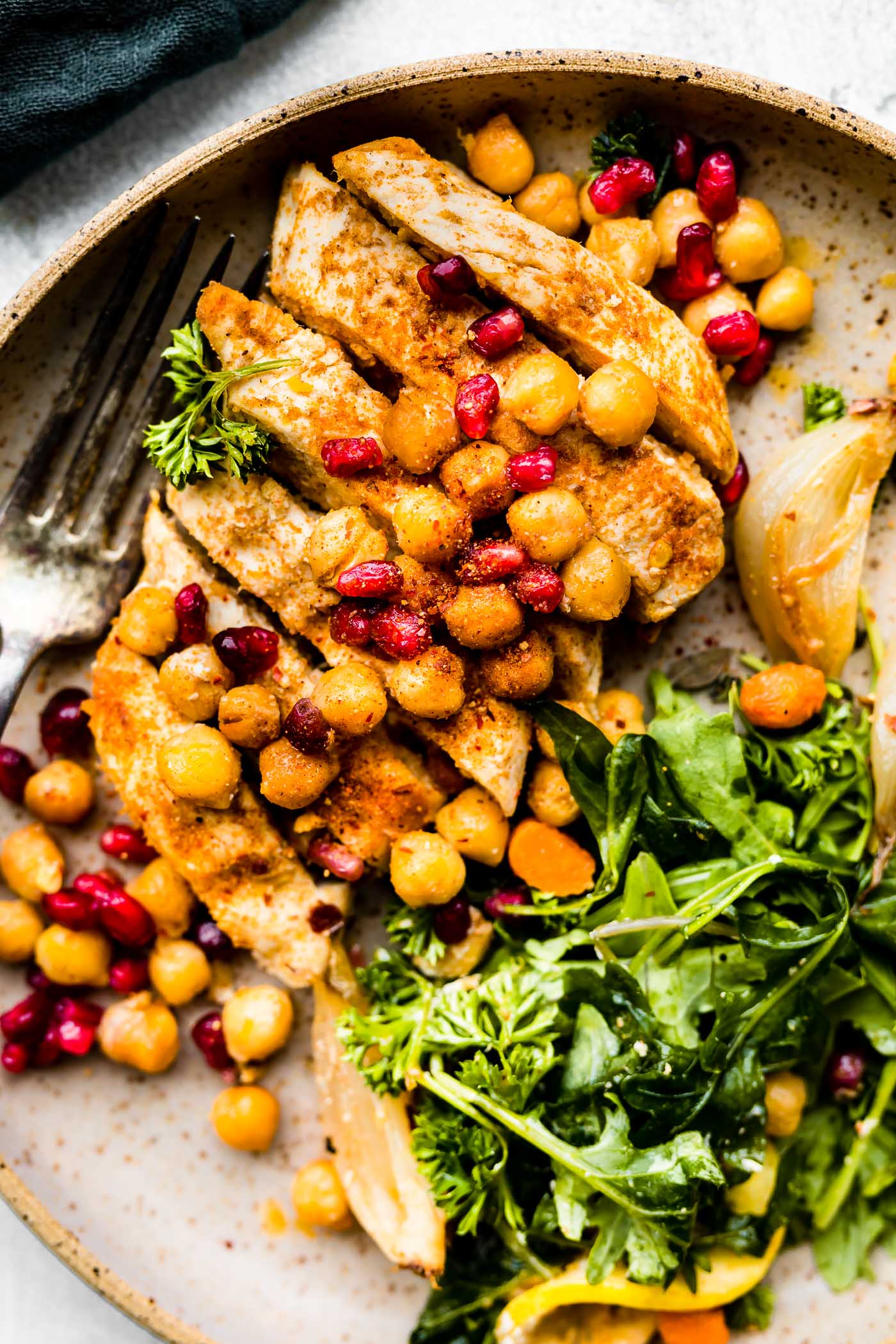 Close up view cumin roasted chicken, sliced topped with roasted chickpeas and pomegranate seeds on plate.