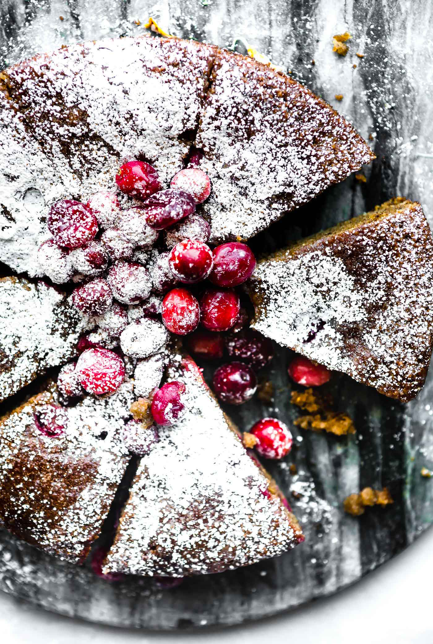 Overhead view cranberry sour cream almond cake cut into slices sprinkled with powered sugar, topped with sugared cranberries.