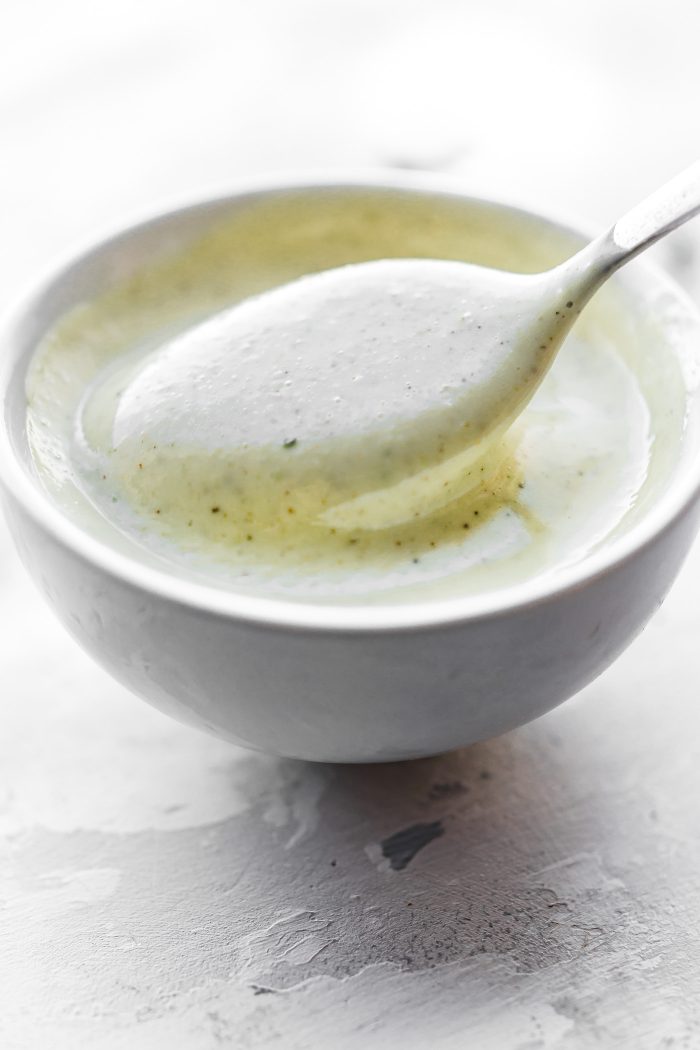 A spoon coming out of creamy homemade ranch dressing in a white bowl.