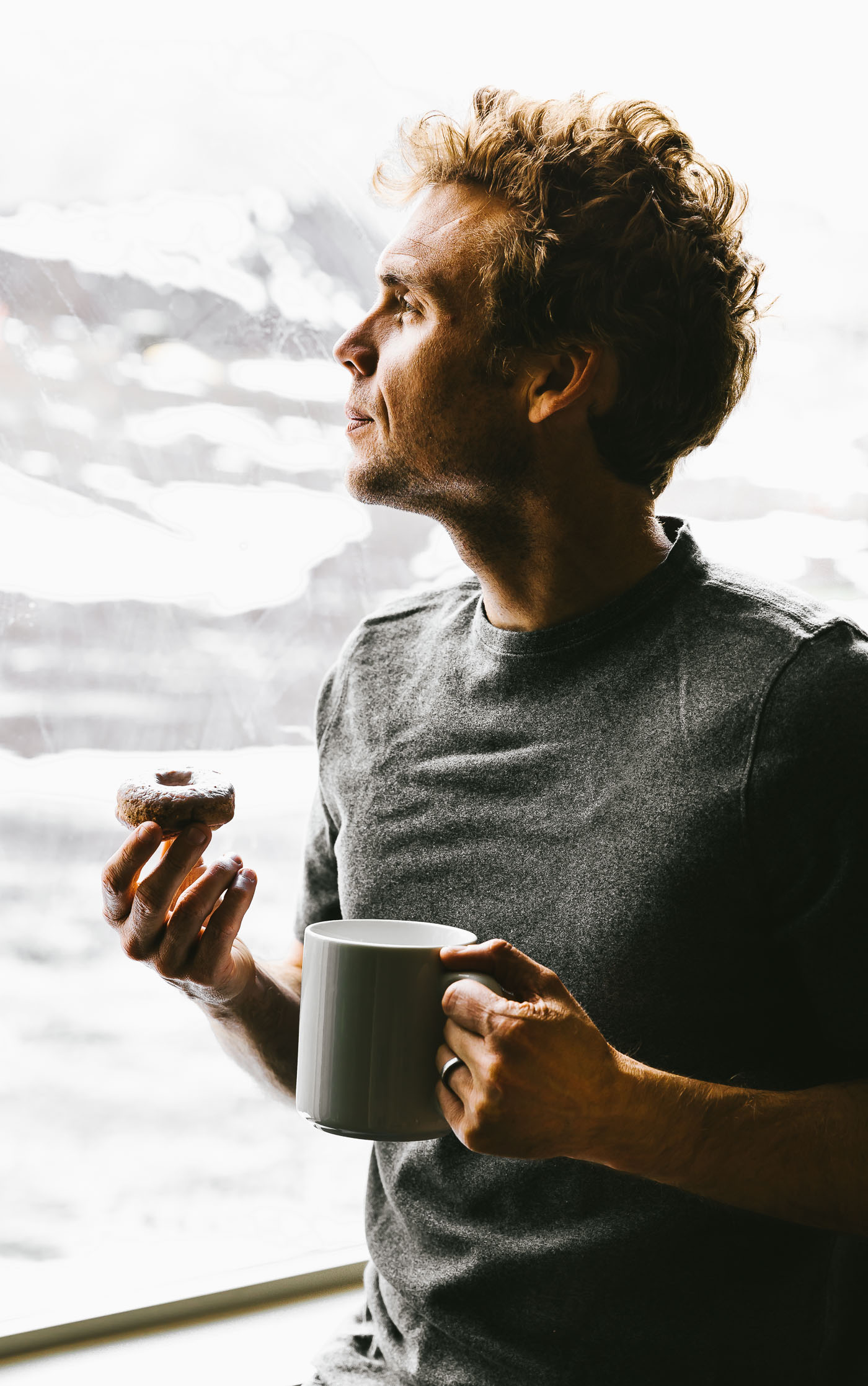 Man standing by bright window holding donut and coffee mug