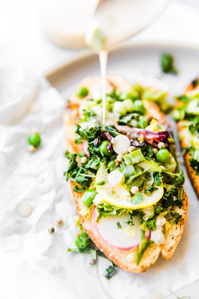 pouring creamy spring onion dressing onto grilled sourdough toast topped with fresh vegetables and herbed cream cheese