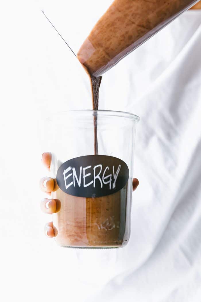A hand holding a glass cup with words 'energy' written on side, a blender cup of chocolate smoothie being poured into glass cup.