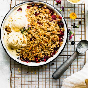 Overhead view metal pie plate filled with coconut berry crisp on cooling rack, melting ice cream on half of crisp